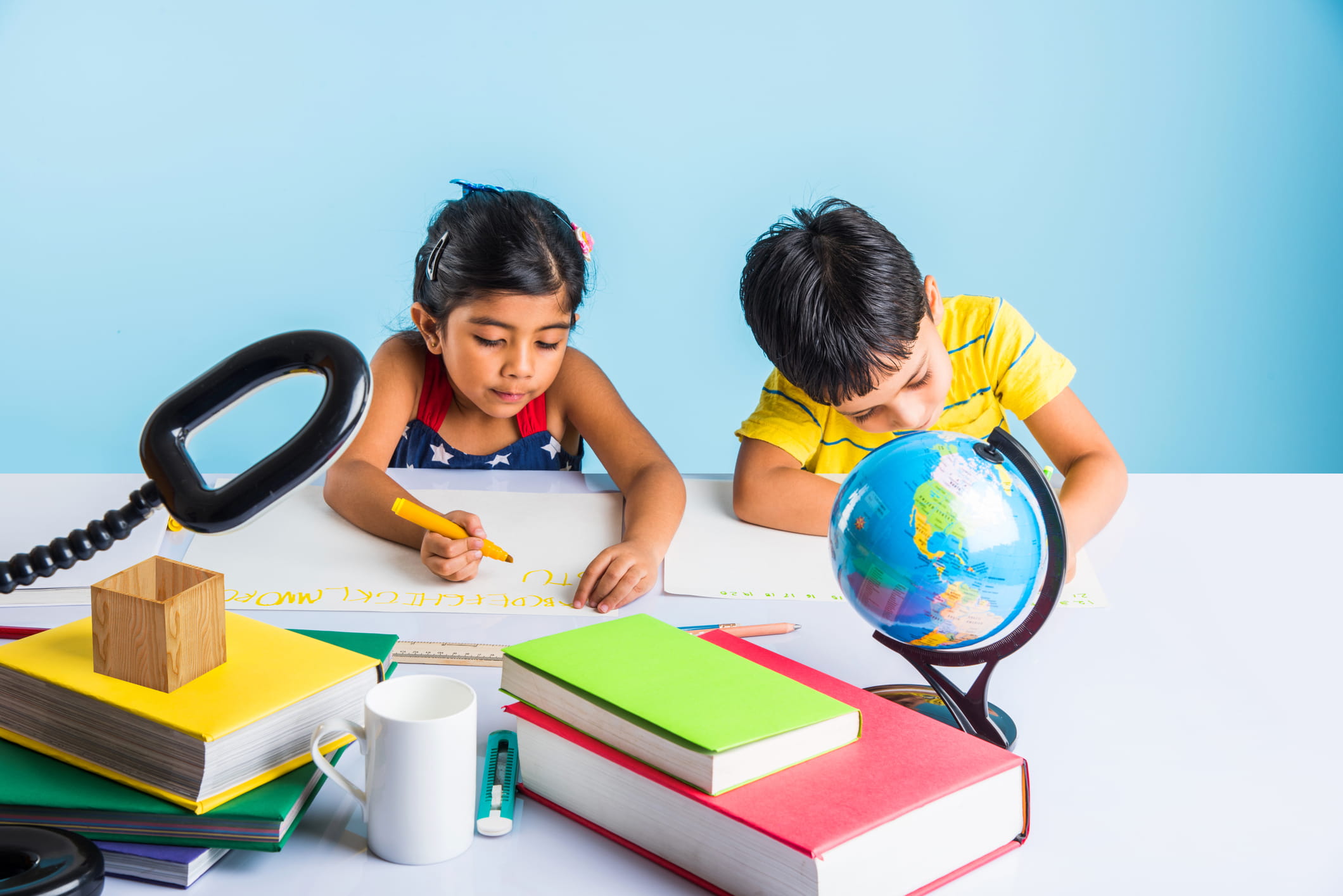 Two young children studying at a desk with books, stationery, and a globe, illustrating early planning for a child’s higher education.