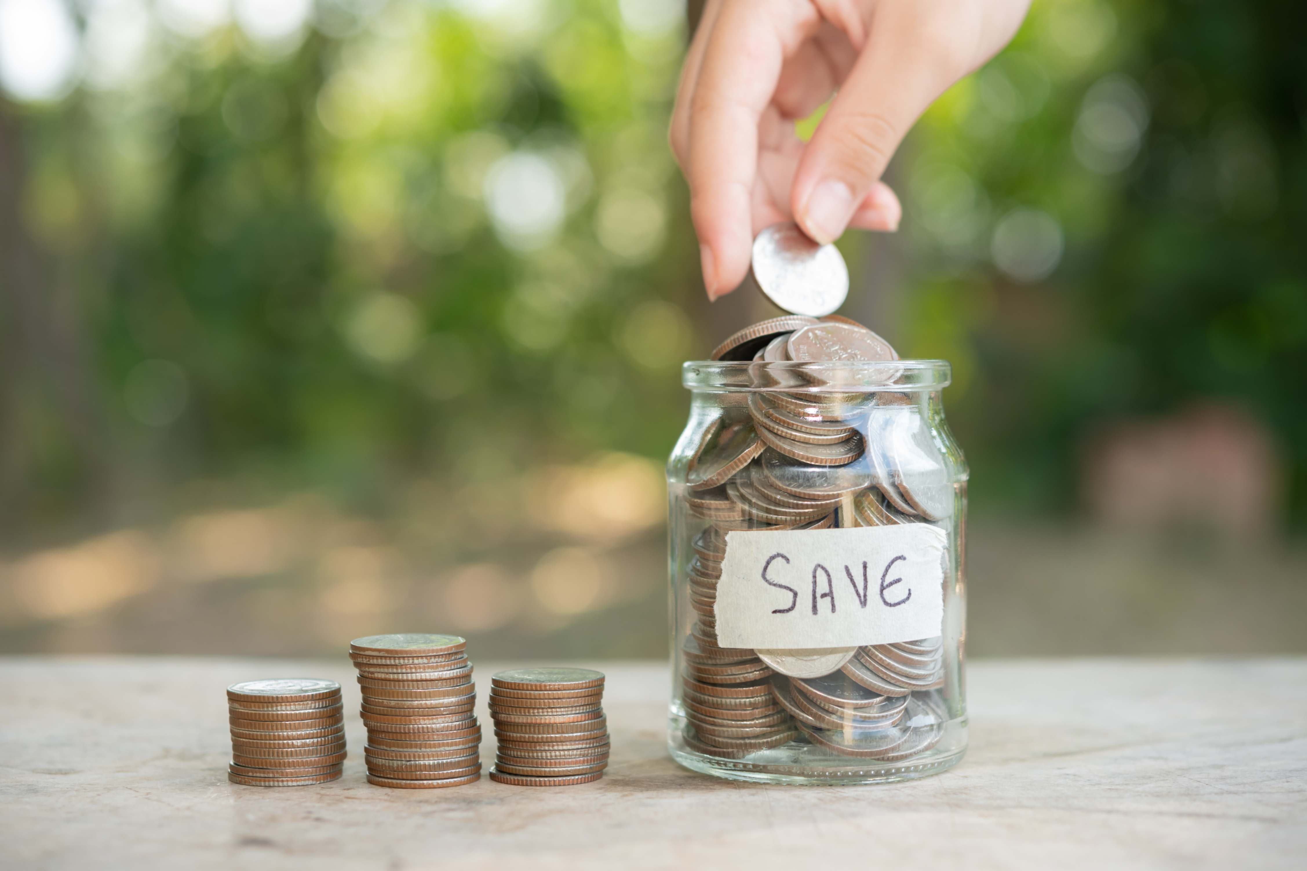 Person placing a coin into a glass jar labelled ‘Save’ beside stacked coins, symbolising budgeting, cash flow management, and saving money to invest toward financial goals.